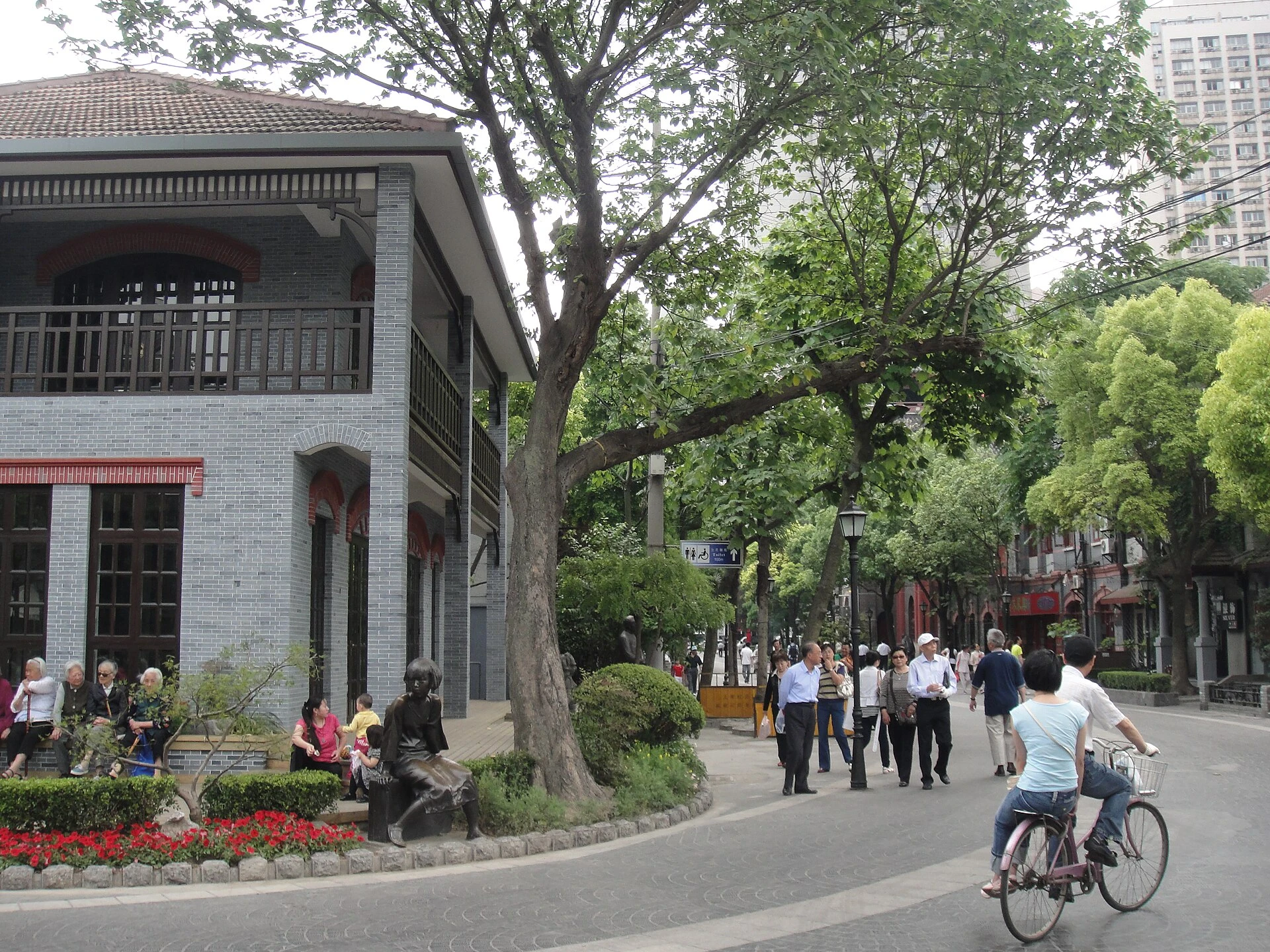 People walking and cycling along Duolun Road beside trees and a literary sculpture